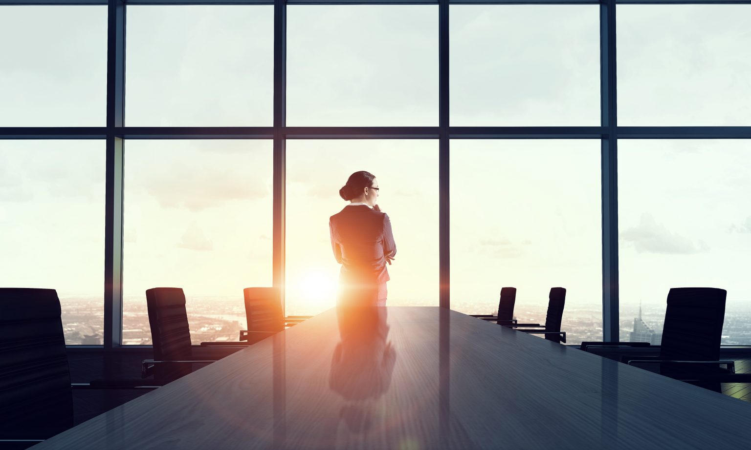 Woman in suit at head of empty boardroom table looking over the city
